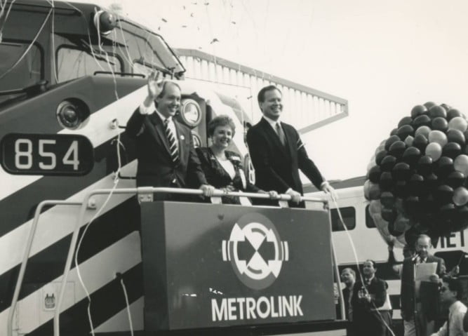 Three people in front of a train with streamers and balloons in the background