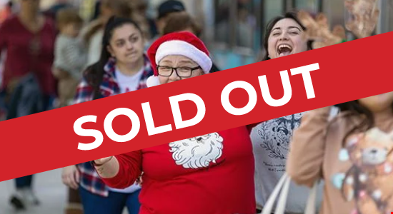 An excited passenger wearing a red sweater with a drawn Santa Claus image while also wearing a classic Santa Hat has two thumbs up as they, and customers around, are boarding the Metrolink Holiday Express Train.